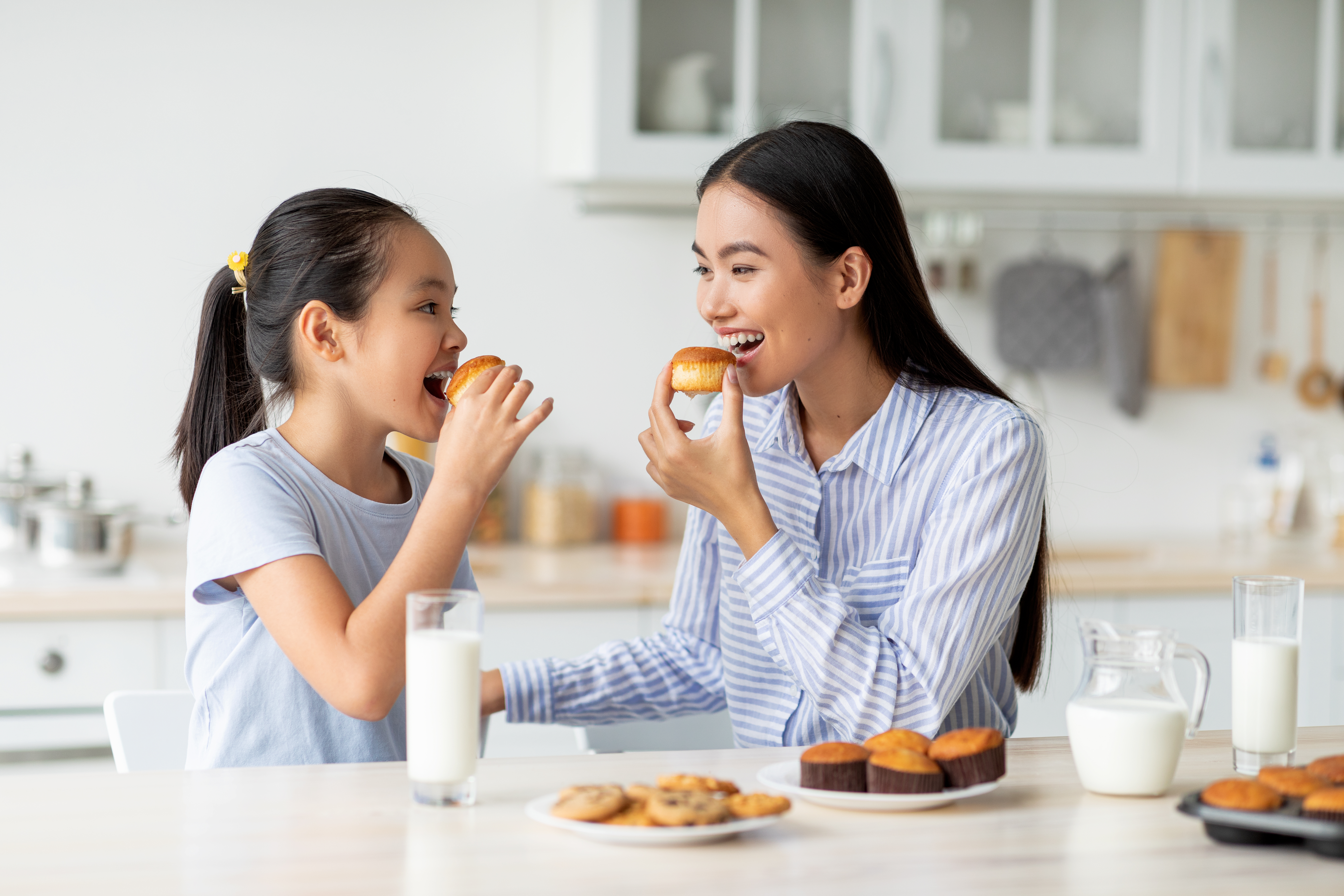 Dessert time for sweet tooth. Happy asian girl and young woman eating cookies and looking at each other, sitting at dinner table in kitchen, enjoying breakfast with milk